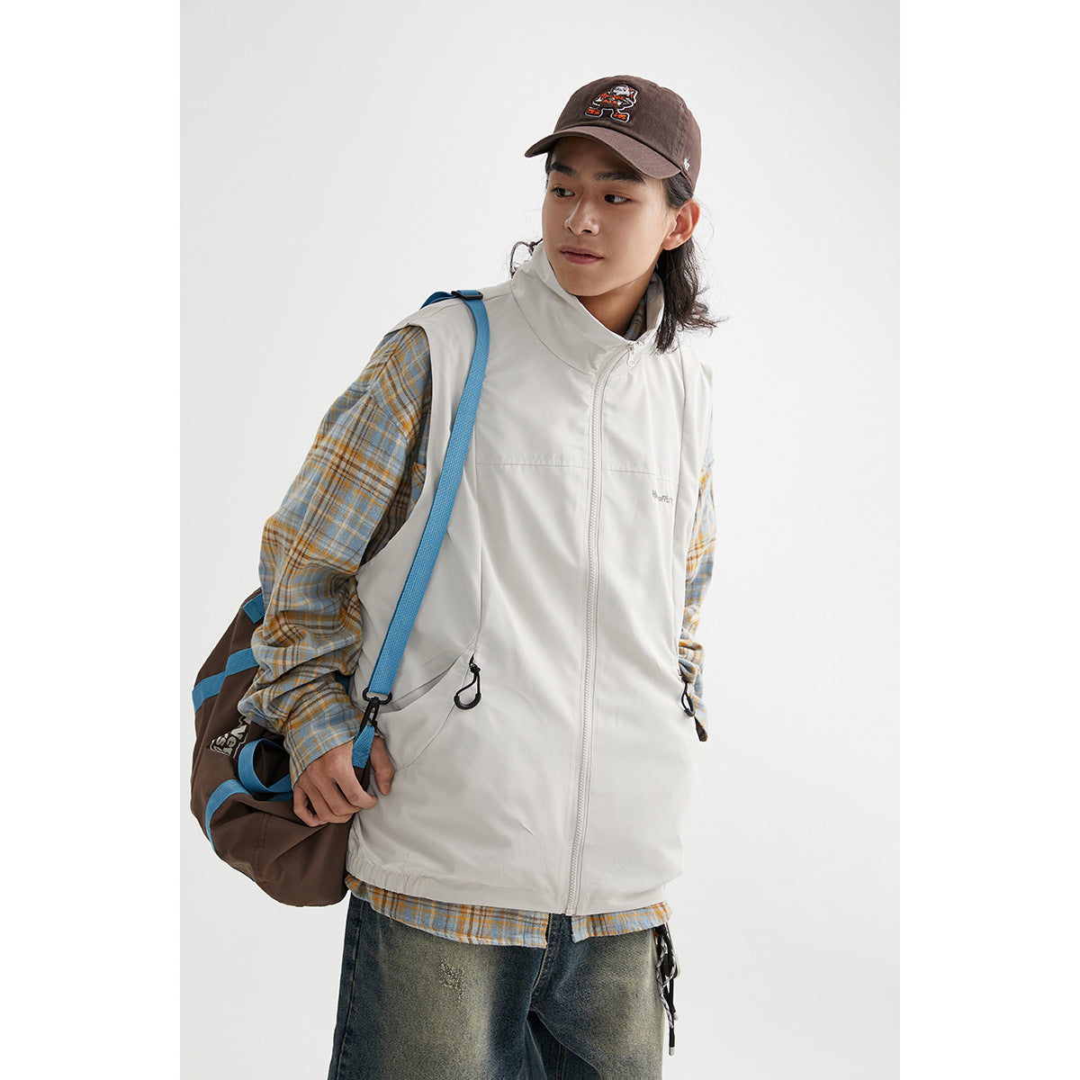 Model wearing a light gray sleeveless zip vest with a high collar and drawcord pockets over a plaid shirt.