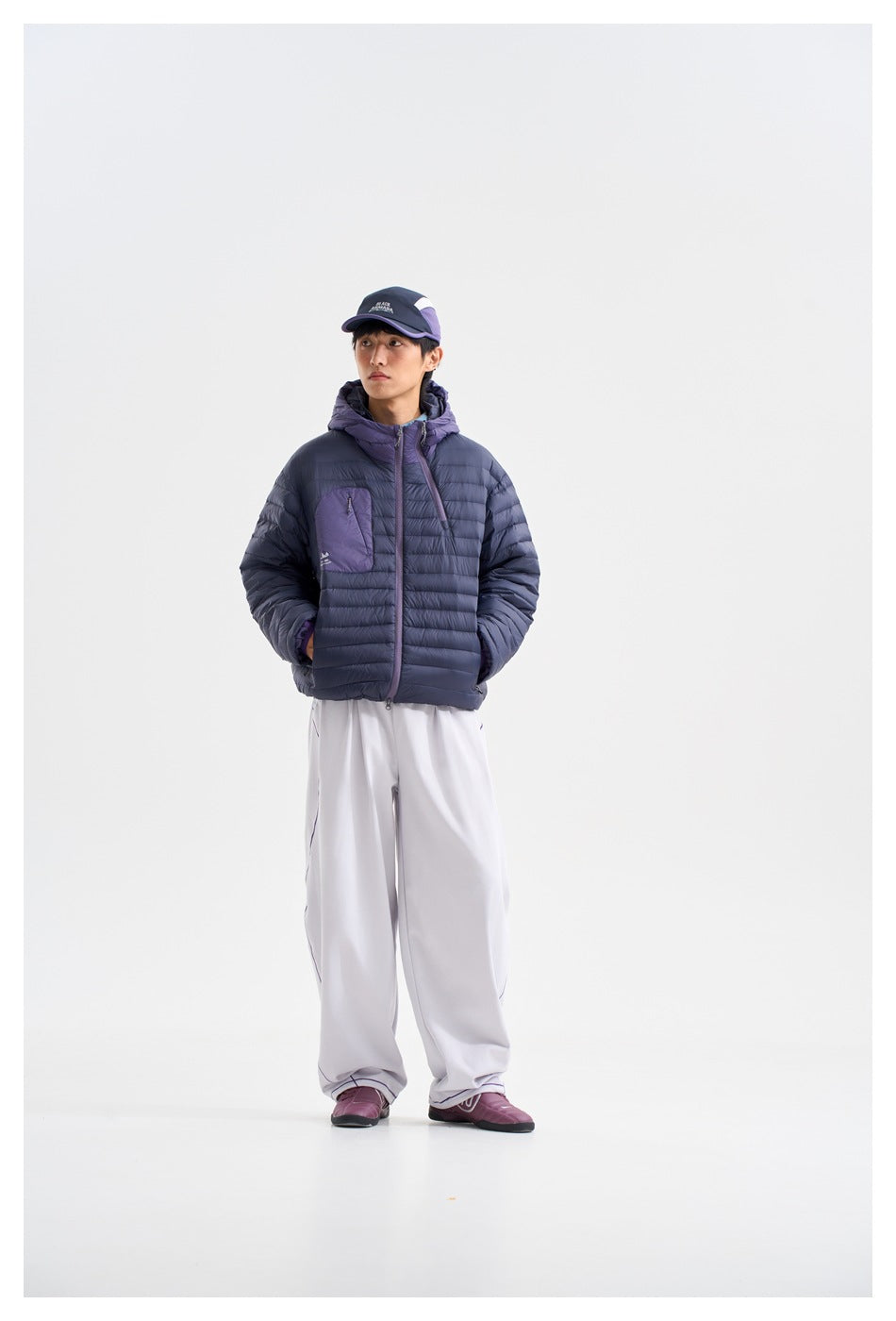 Model wearing a navy hooded puffer jacket with a purple chest pocket and loose white pants.