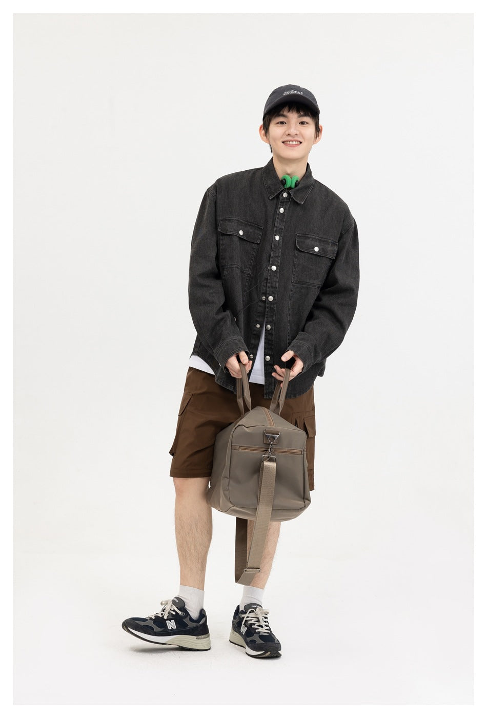 Smiling model wearing the washed black shirt over a white tee with brown shorts, holding a taupe duffel bag