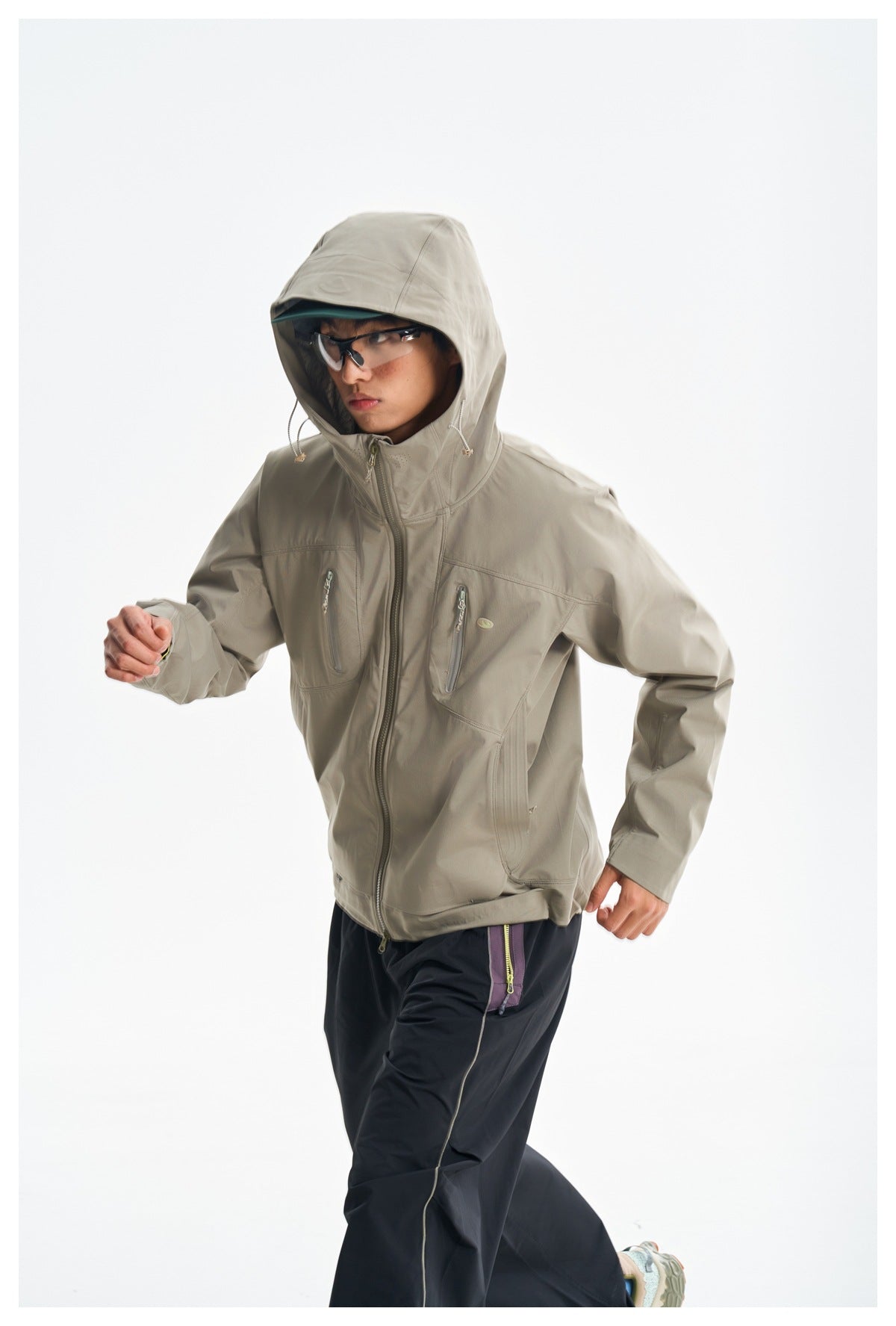 Model wearing a light gray-beige hooded jacket with zip pockets and dark pants, shown in a dynamic pose.