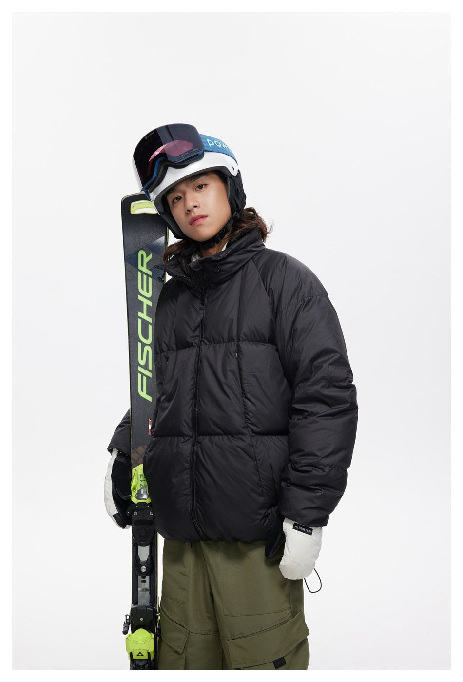 Model wearing a black quilted puffer jacket with ski helmet and gloves, holding skis.