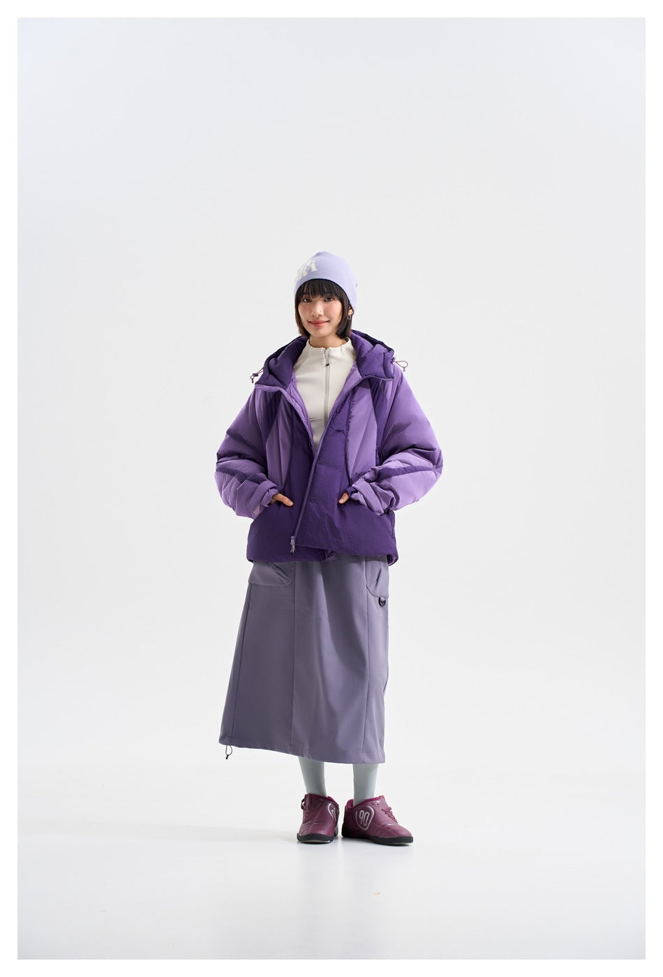 Model wears an open purple hooded jacket over a light zip top, with a long gray-purple skirt.