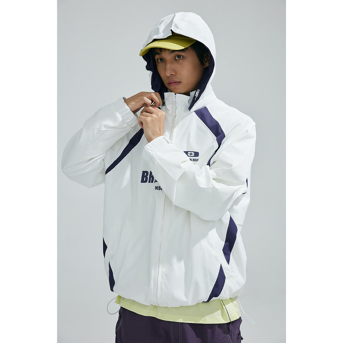 Model wearing a white hooded zip jacket with navy panels and printed chest graphics, paired with purple shorts.