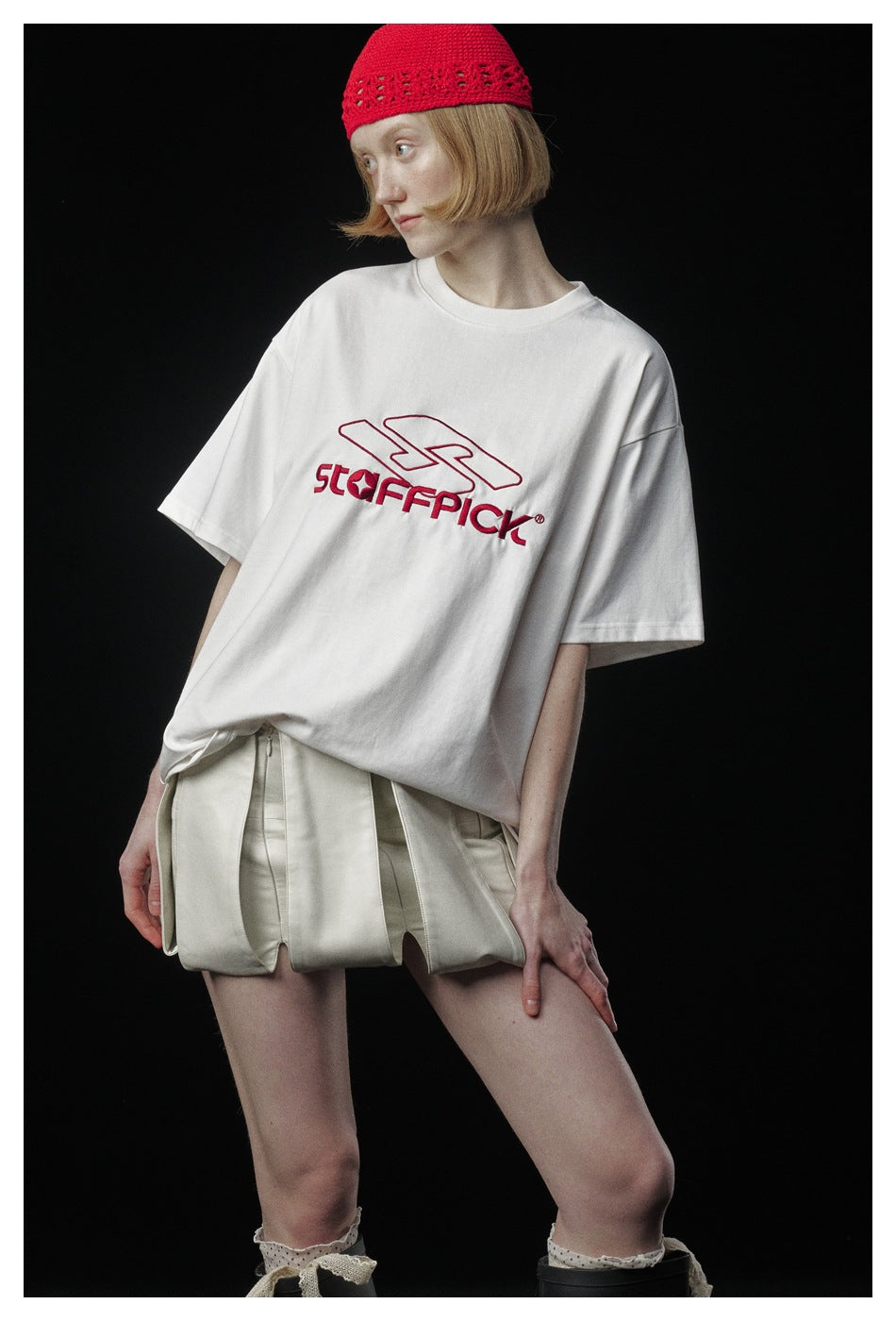 Model wearing an oversized white T-shirt with red chest lettering, cream shorts, and a red knit cap.