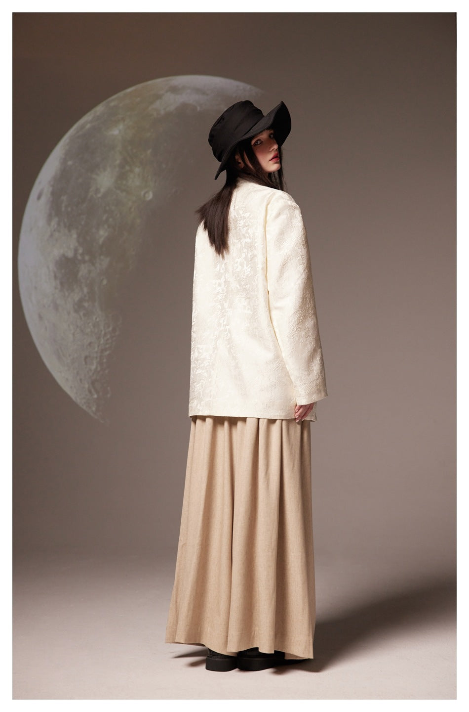 Back view of a model wearing an ivory floral-textured jacket, beige ankle-length skirt, and black hat.
