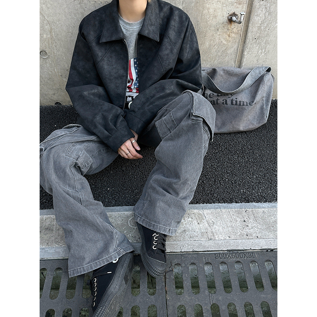 Seated model wearing a dark charcoal zip-front jacket and loose gray cargo pants.