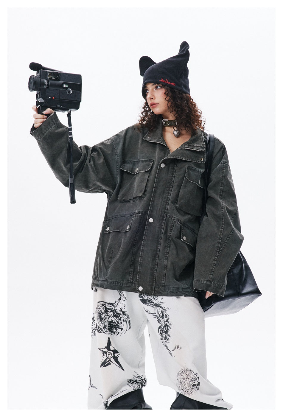 Model in a charcoal oversized utility jacket and white graphic trousers, holding a video camera.