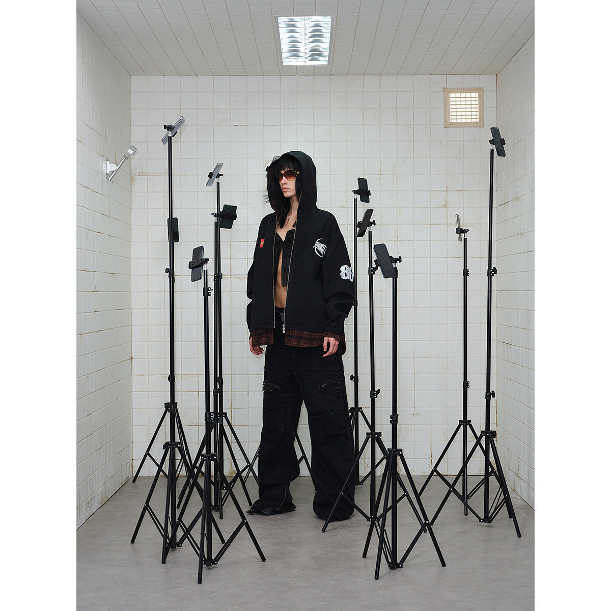 Model wears a black graphic hooded zip jacket and wide distressed black pants, surrounded by equipment stands.