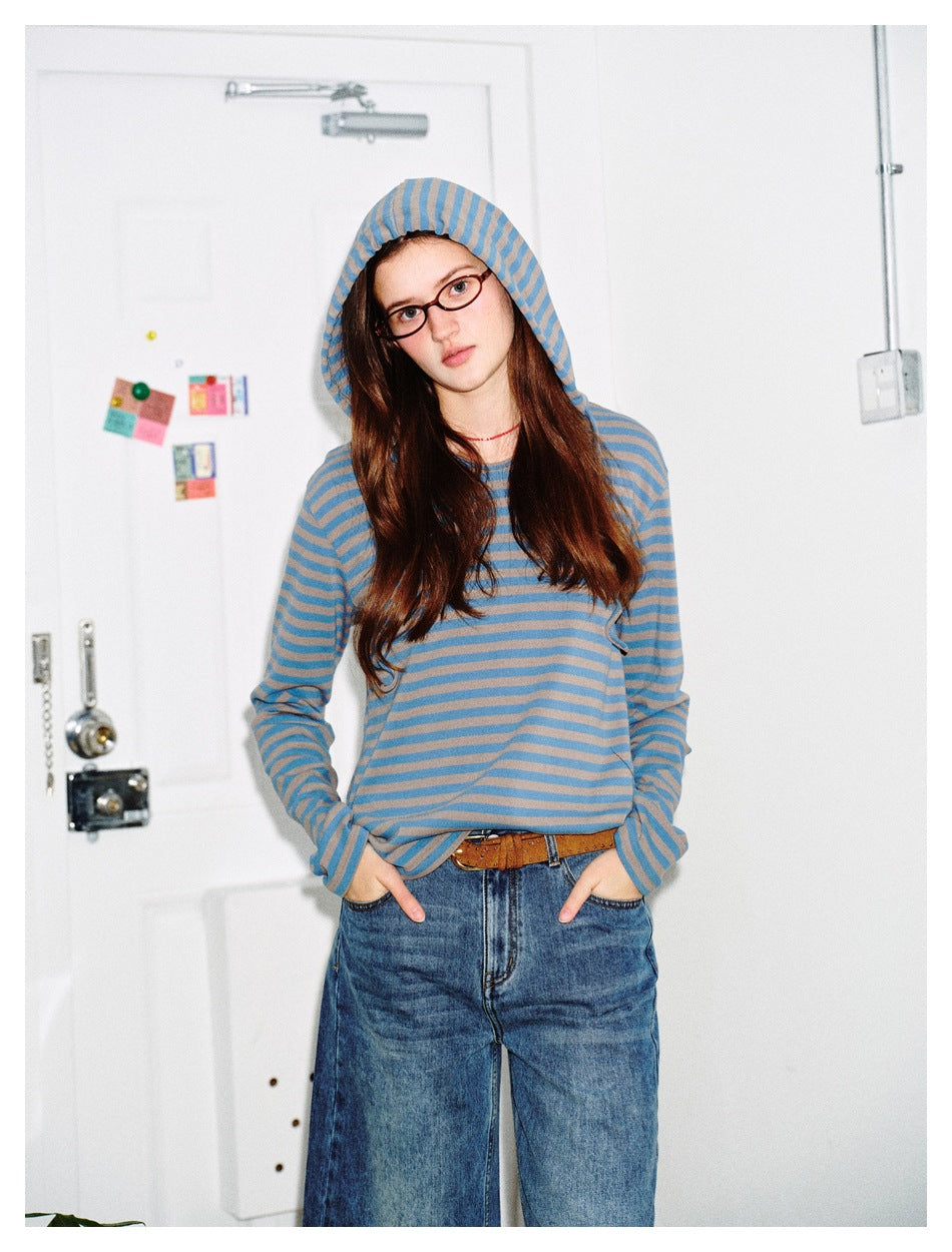 Model wearing a blue and taupe striped hooded long-sleeve top with jeans, hands in pockets.