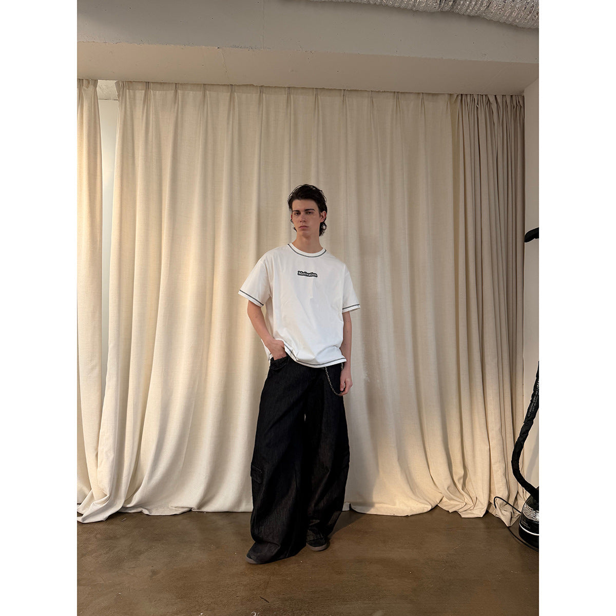Model wearing a white T-shirt with black contrast stitching and a small chest graphic, paired with loose black jeans.