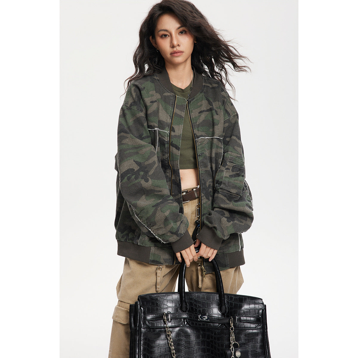 Model wearing an open, oversized dark green camouflage bomber jacket with contrast seam details and a zippered sleeve pocket.