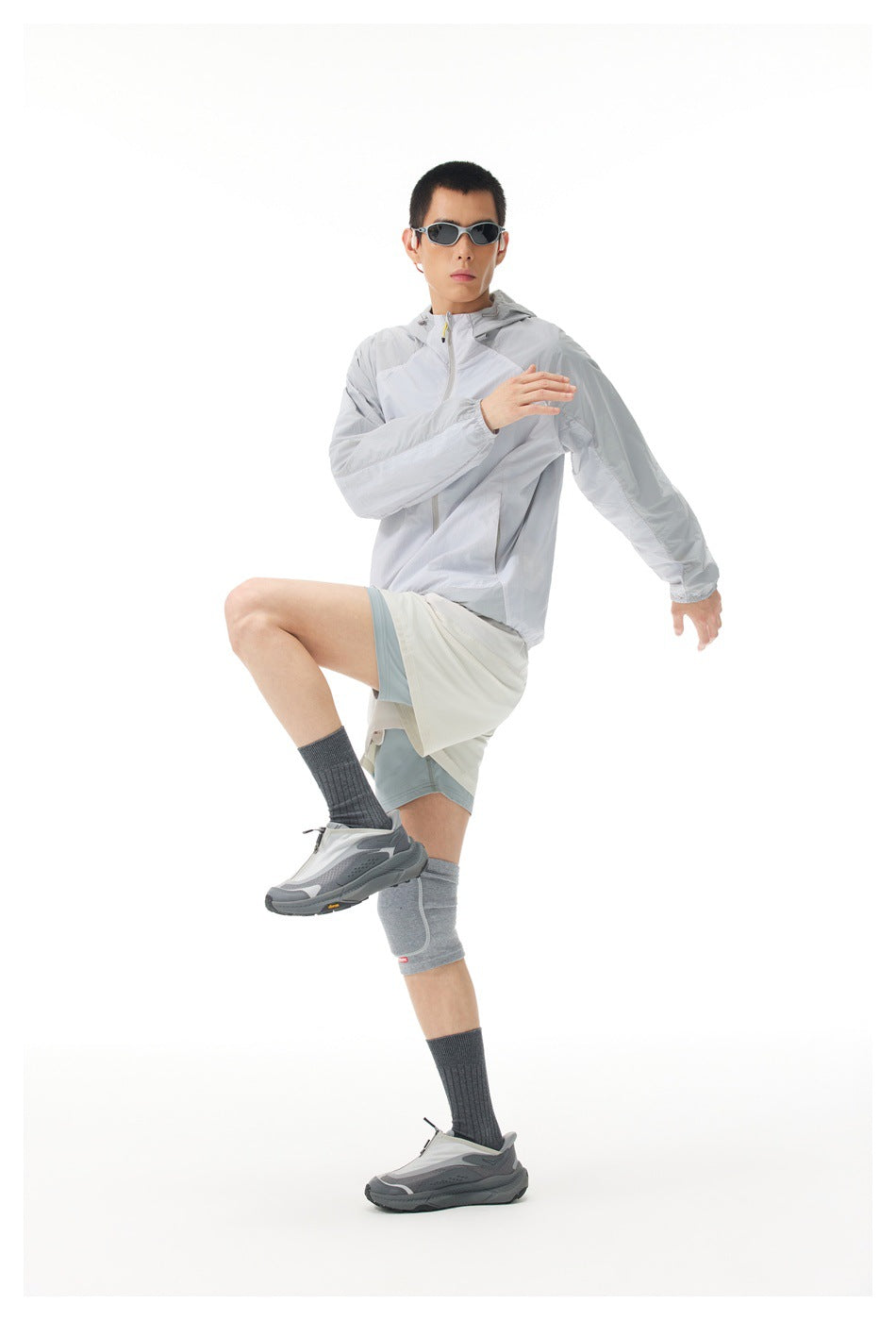 Model in a light gray hooded zip jacket and layered shorts, posed balancing with one knee raised.