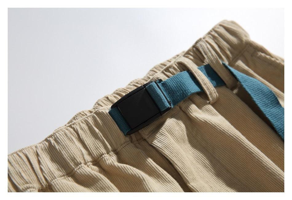 Close-up of beige ribbed trousers with an elastic waistband and a teal webbing belt with black buckle.