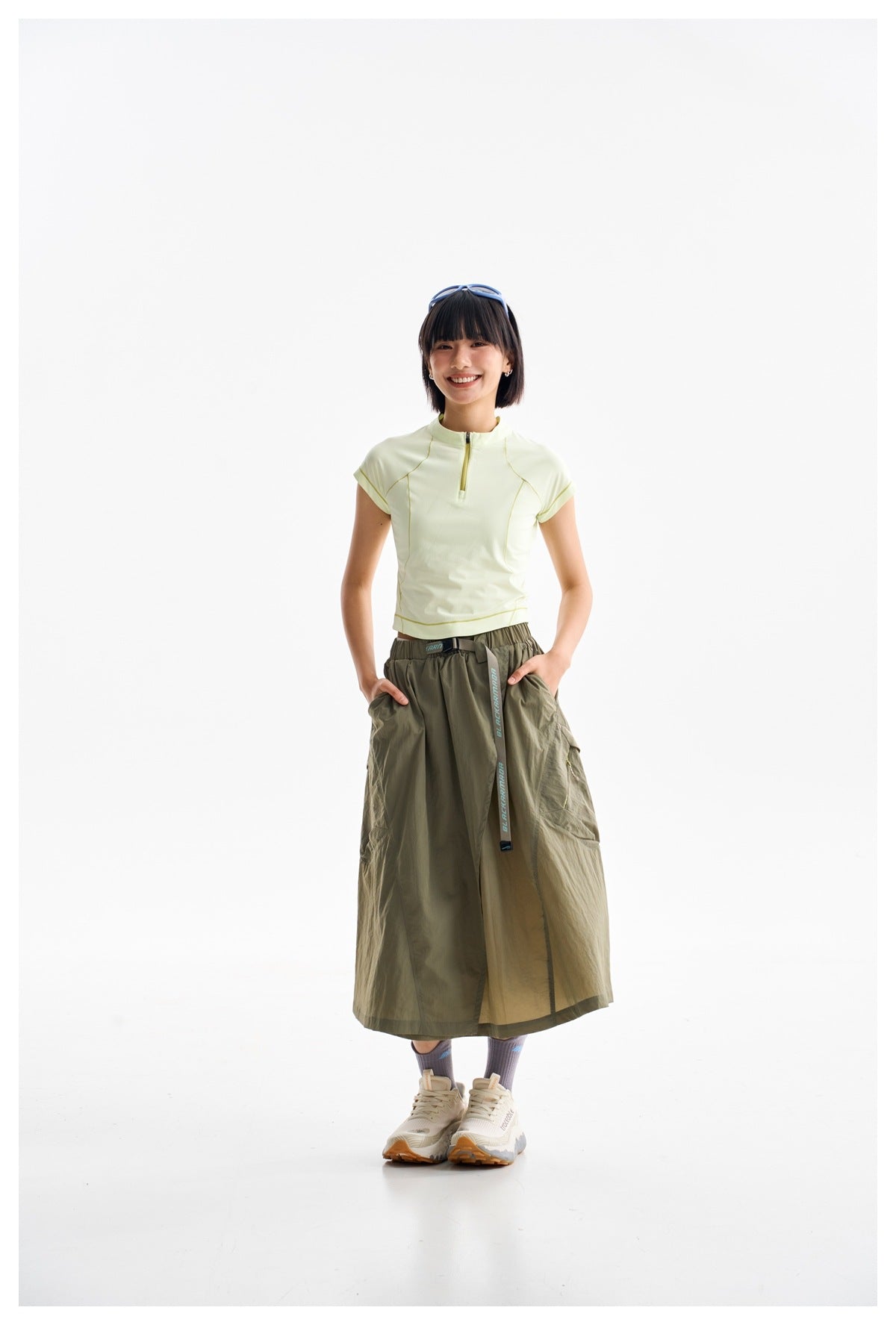 Full-length model wearing a pale yellow zip-neck top, olive wrap-style midi skirt, socks and sneakers.