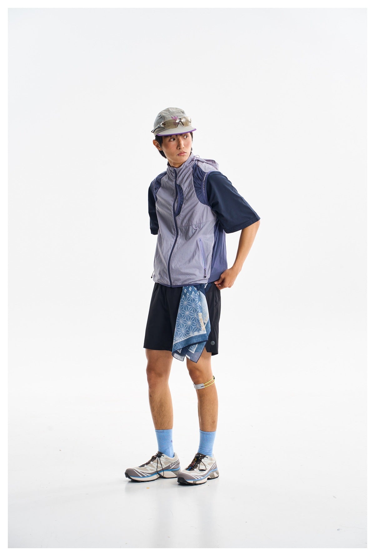 Three-quarter view of a man wearing a light gray hooded vest over a navy shirt with navy shorts and a gray cap.