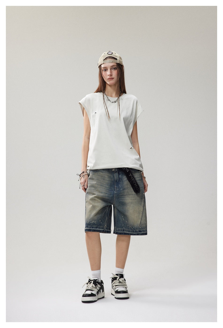 Model wearing a light beige sleeveless distressed top with denim shorts, cap and sneakers, front view