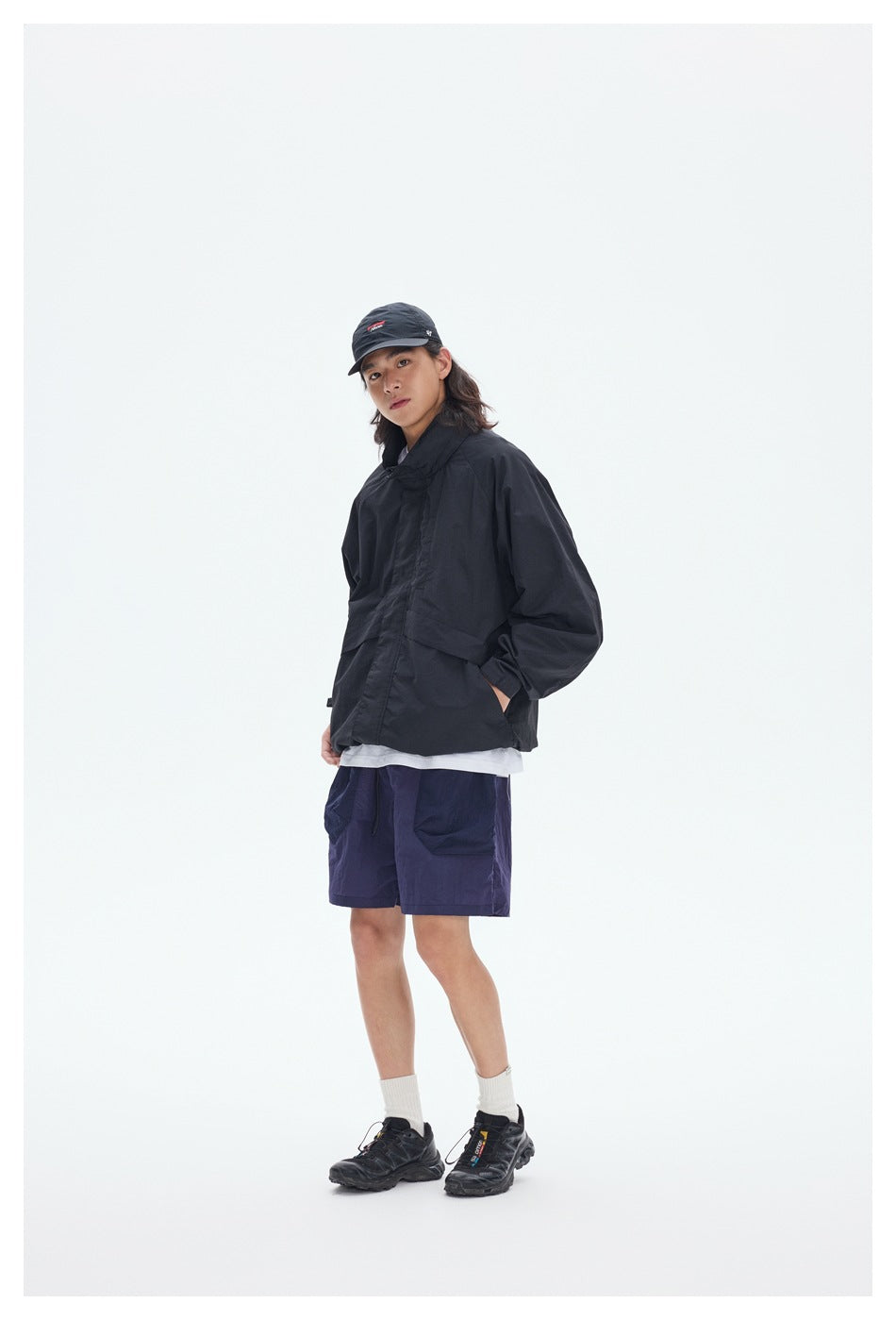 Model wearing a black oversized jacket with navy shorts, a black cap, white socks and dark sneakers.