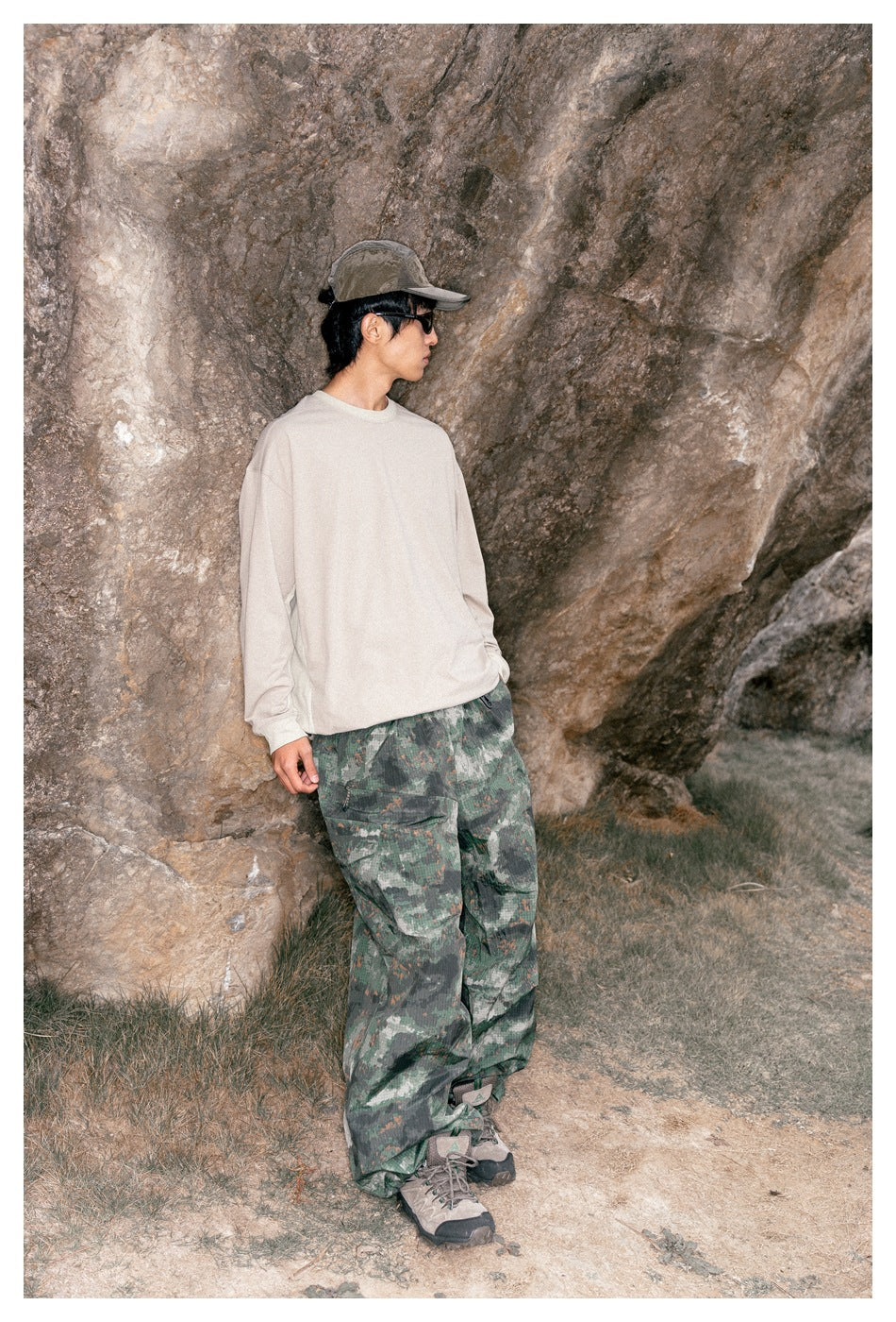 Model wearing a loose cream long-sleeve shirt and wide green camouflage cargo pants beside a rocky wall.