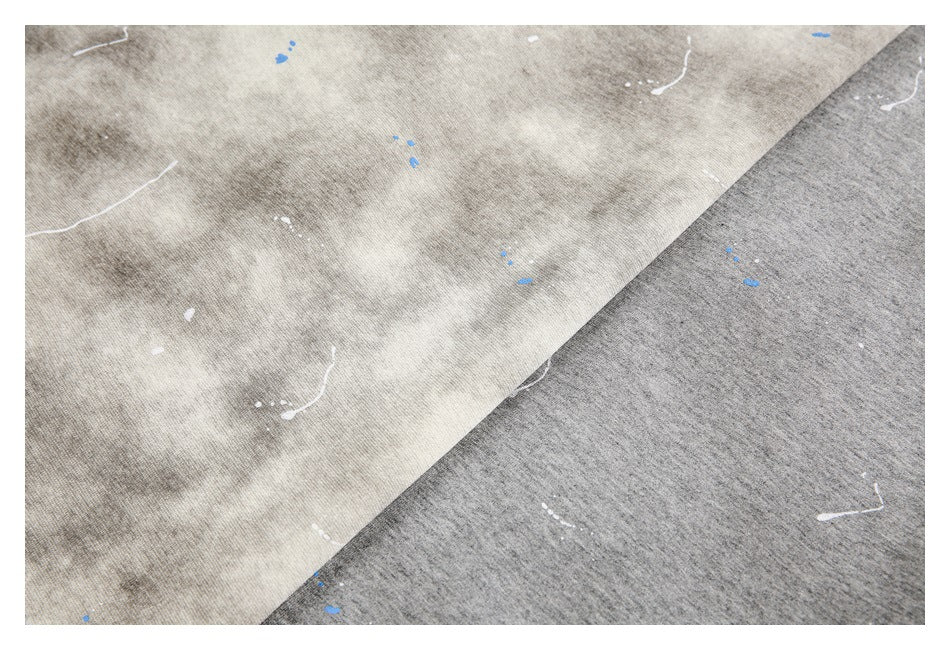 Close-up of beige-gray and charcoal-gray shirt fabric with cloudy dye patterns and scattered white and blue marks.