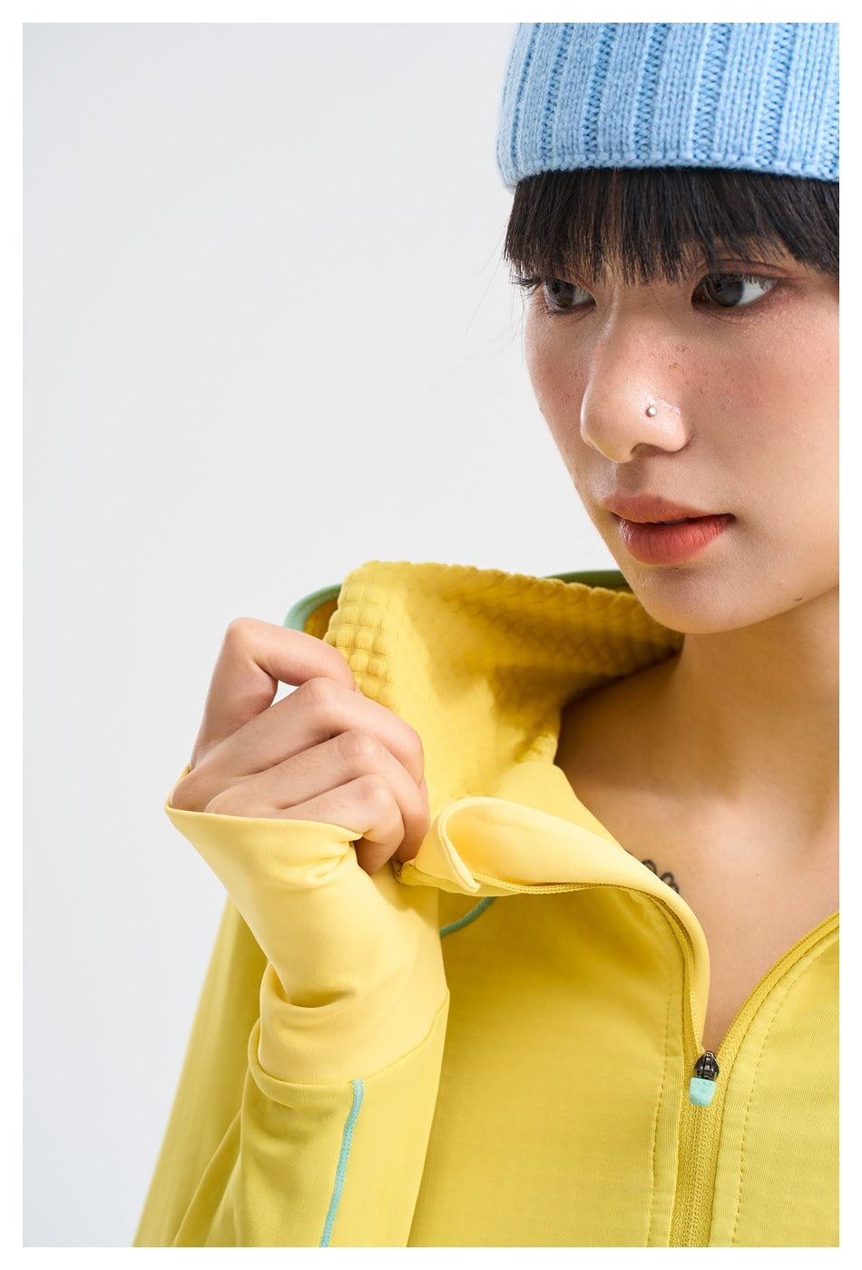 Close-up of a model wearing a yellow zip-up jacket, showing its hood, zipper, and extended thumbhole cuff.