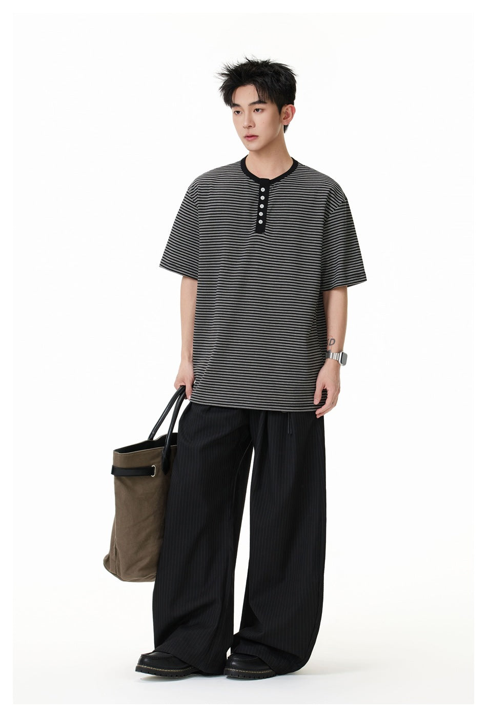Model standing in a black and gray striped henley, carrying a brown tote bag.