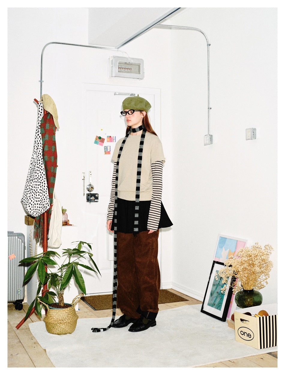 Three-quarter view of a model in a beige layered top, black skirt, brown trousers, green beret and long striped scarf.