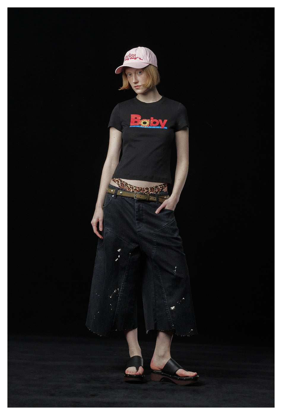 Model wearing a black graphic T-shirt with wide cropped black jeans and a pale pink cap.