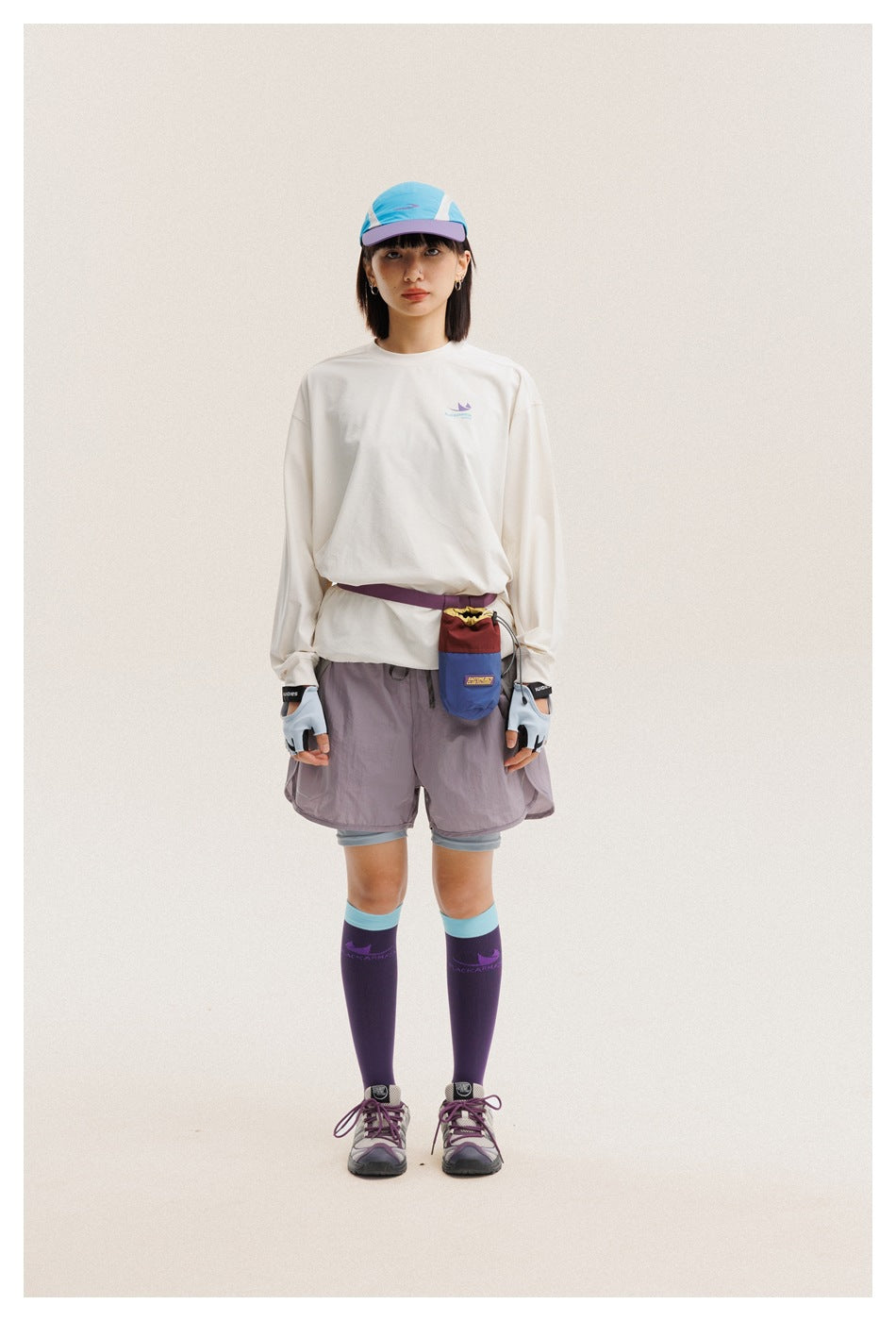 Front view of a woman wearing an off-white long-sleeve shirt, mauve shorts, a colorful cap, gloves, and purple knee socks.