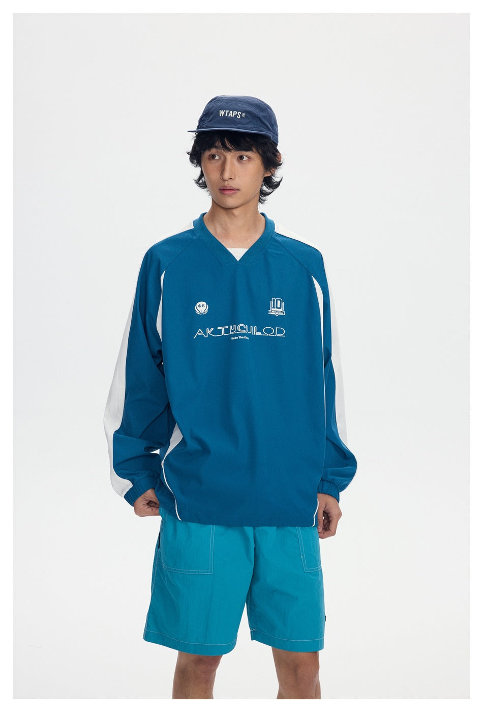 Closer front view of a model wearing a blue-green V-neck pullover with white sleeve panels and teal shorts.