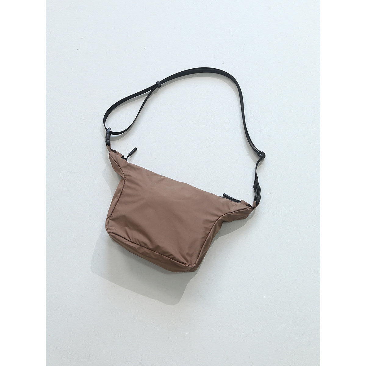 Brown crossbody bag with black adjustable strap, buckles and zipper pull on a light background