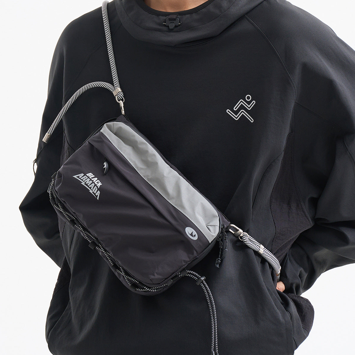 A person wears a black crossbody utility bag with a gray panel, cord strap and white Black Armada lettering.