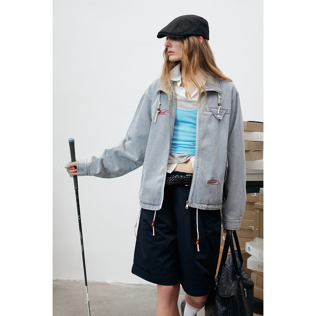 Model wearing a blue-and-white pinstriped zip jacket with red embroidery, styled with a black cap and navy shorts.