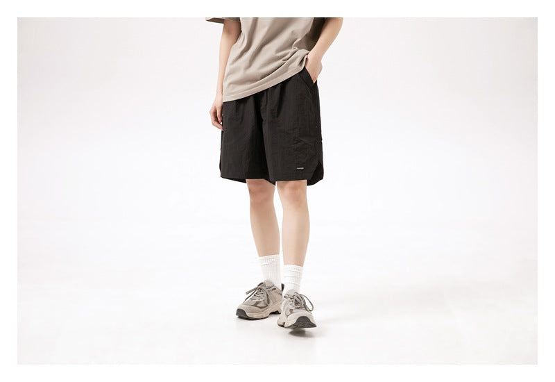 Model wearing loose black shorts, shown from a slight front angle with one hand in a pocket.