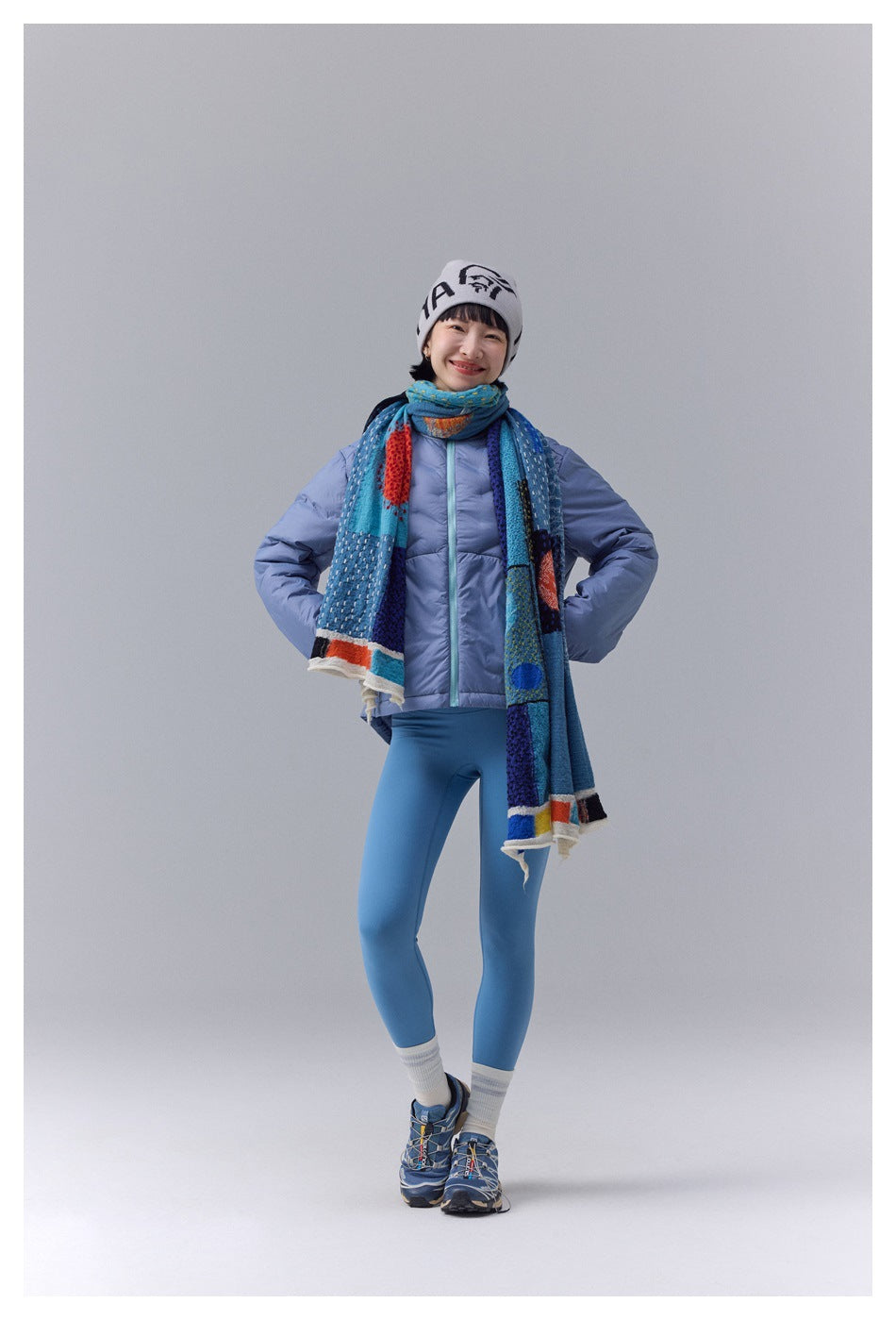 Model wearing a light blue quilted jacket with a turquoise front zipper, paired with a multicolored scarf.