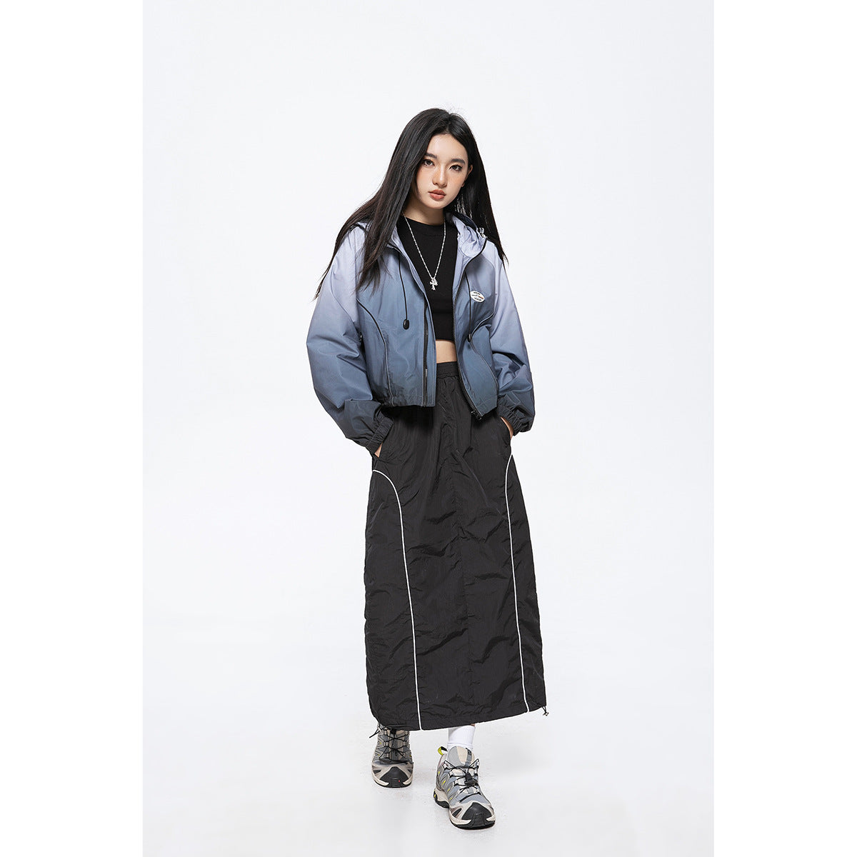 Model wearing a blue-gray hooded zip jacket and a long black skirt with white piping, shown from the front.