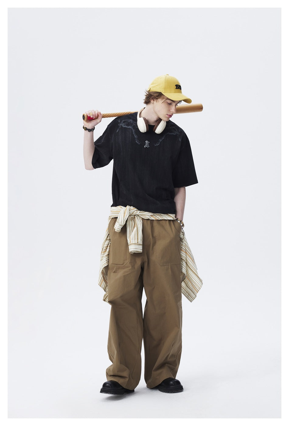 Full-length model wearing an oversized black graphic T-shirt with olive cargo trousers and a yellow cap.