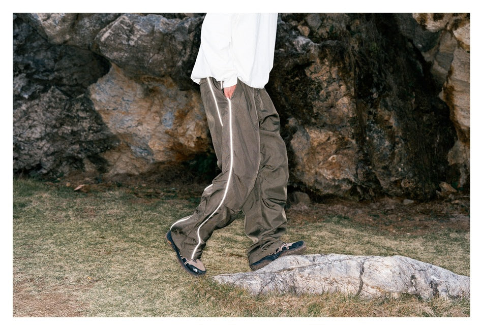 Side view of a model wearing loose taupe-gray pants with bright reflective piping down the leg.