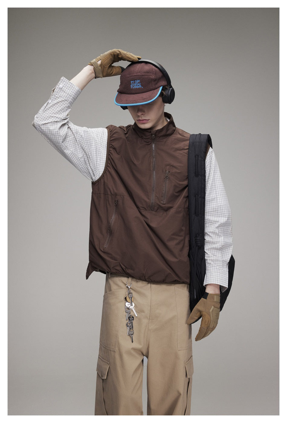 Model wearing a dark brown zip-front vest over a checked shirt and beige cargo pants, one gloved hand raised.
