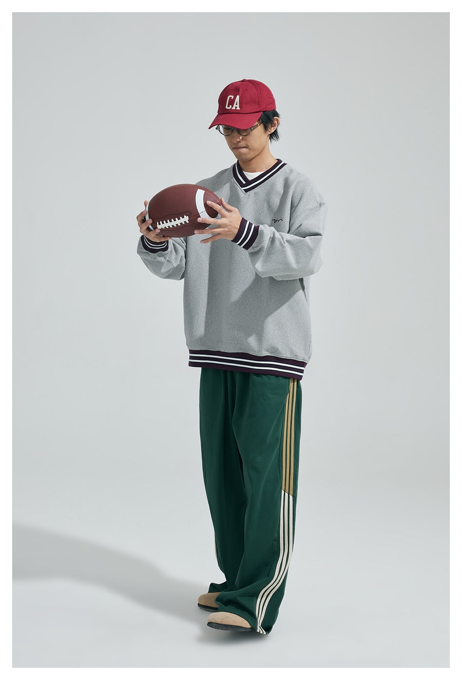 Model wearing a light gray V-neck sweatshirt with dark striped trim and green track pants, holding a football.