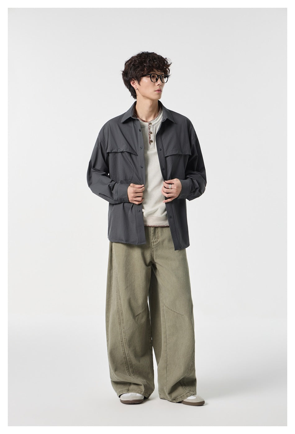 Model wearing a charcoal gray button-front overshirt with flap chest pockets and loose olive-gray trousers.