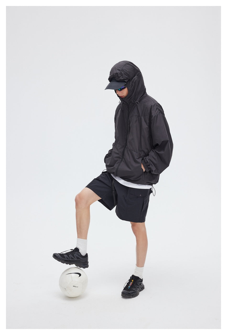 Model wearing a charcoal hooded zip jacket and black cargo shorts, posed with one foot on a soccer ball.