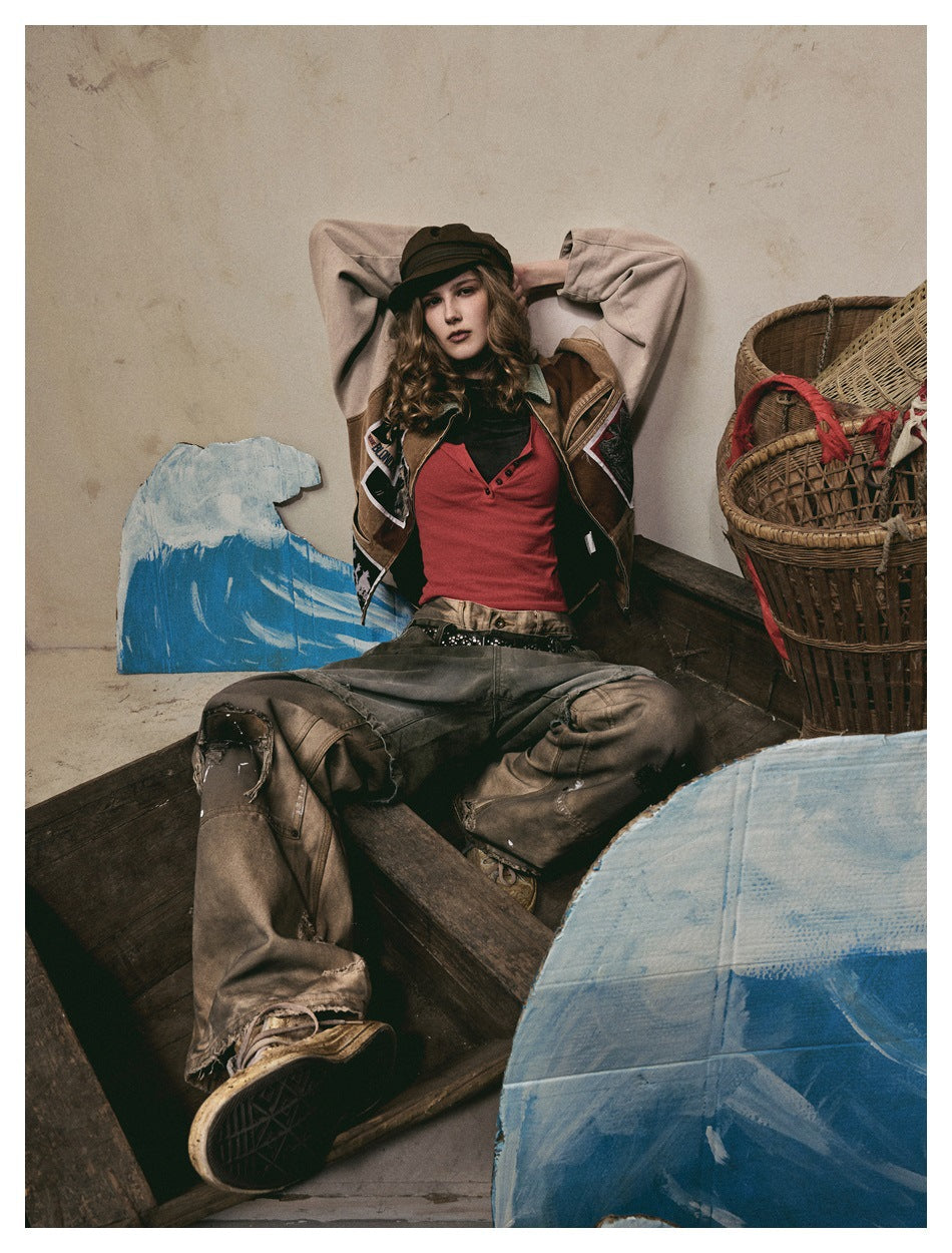 Model seated in a brown patchwork jacket with pale sleeves, red top, dark cap, and loose gray-brown pants.
