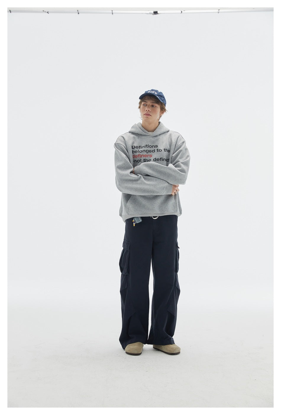 Model in a heather gray printed hoodie with arms crossed, wearing a navy cap and dark cargo pants