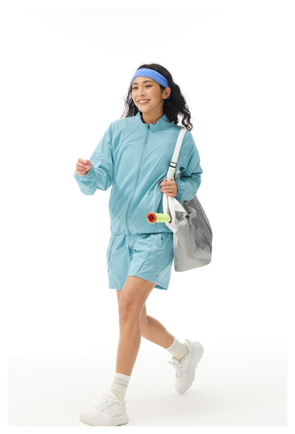 Model in a light blue zip jacket and shorts with a blue headband, carrying a white tote bag.