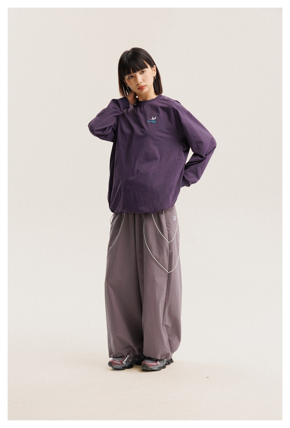 Model wearing an oversized purple long-sleeve shirt with a small chest logo and loose gray-mauve pants.