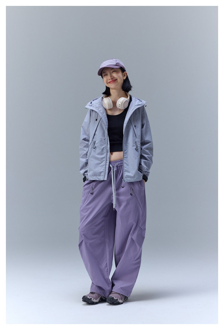 Model wearing a light gray hooded jacket and loose lavender pants, with a lavender cap and headphones.