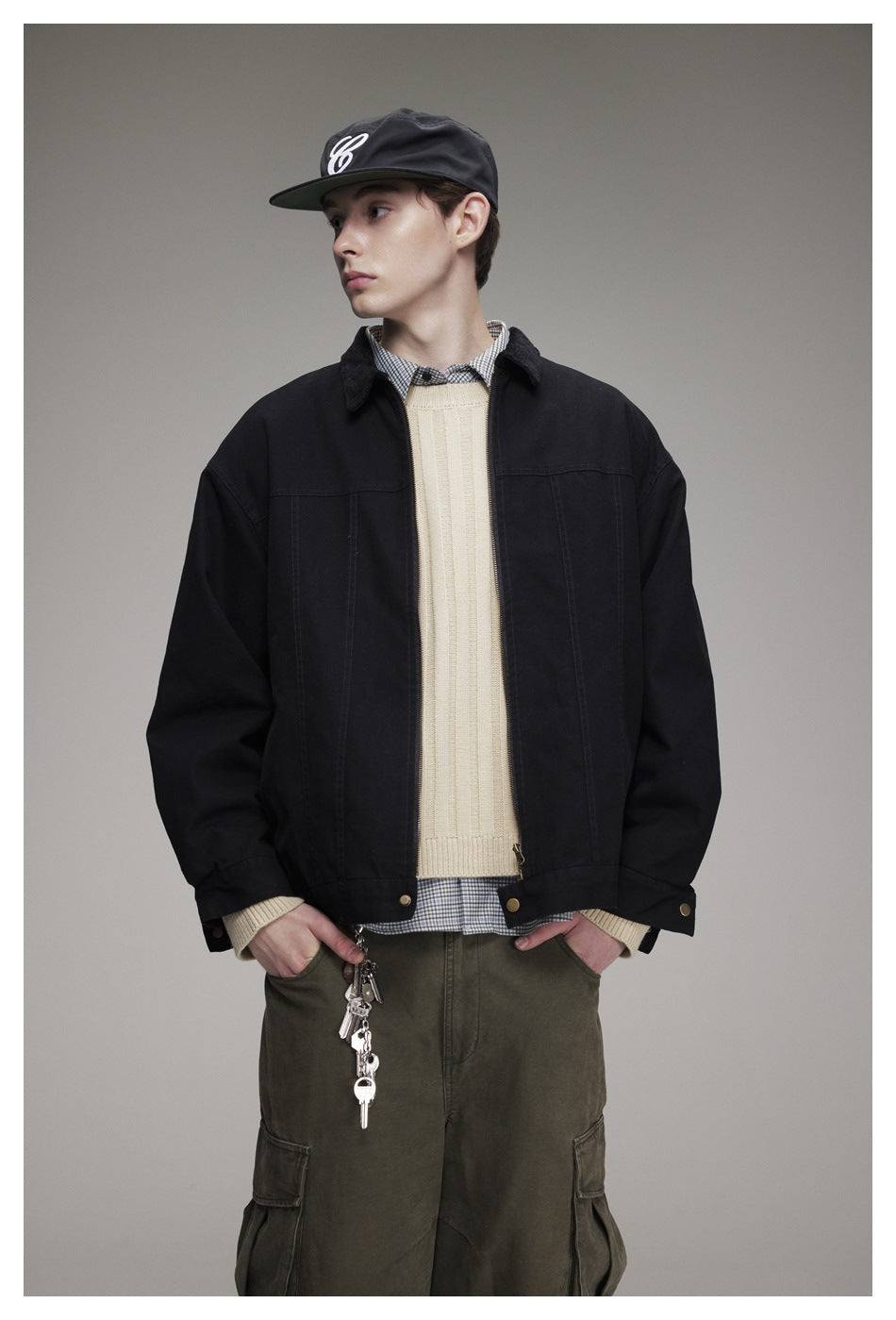 Model wearing a black snap-front jacket over a cream sweater and checked shirt, with olive cargo pants.