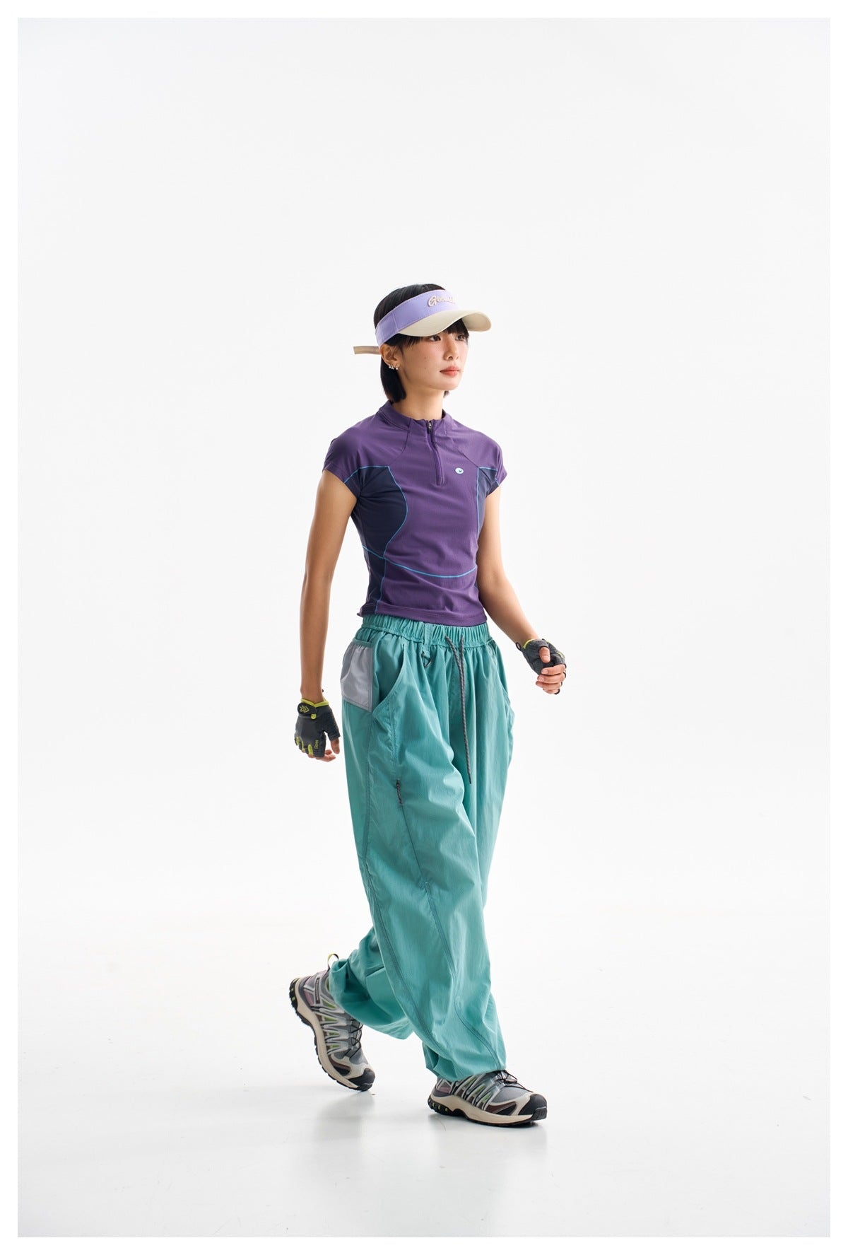 Model walking in a purple zip-neck athletic top and loose teal pants, wearing a lavender visor.