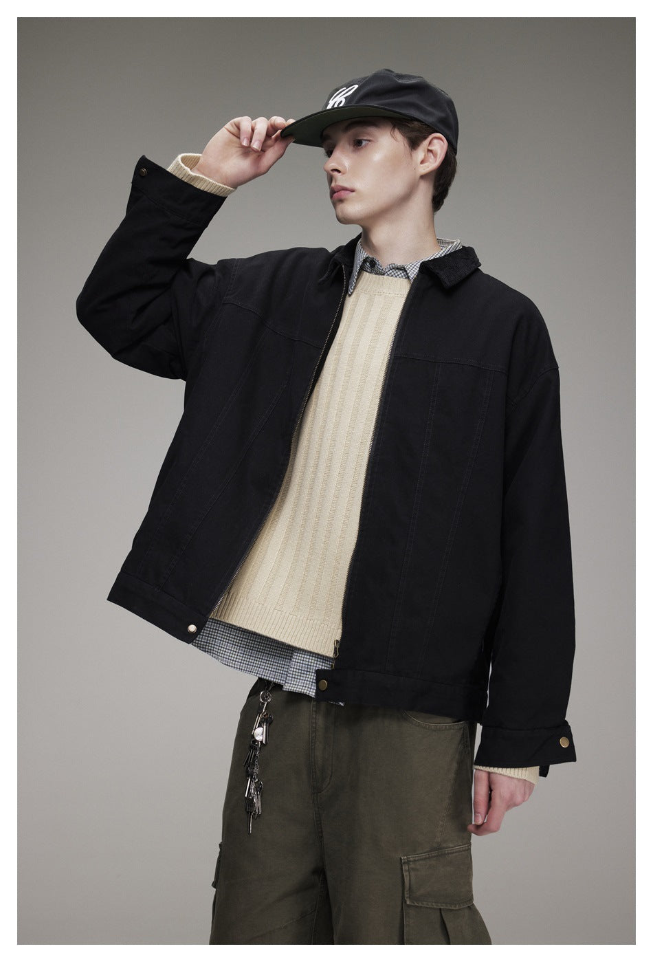 Model wearing a black snap-front jacket over a cream sweater and checked shirt, with olive cargo pants.