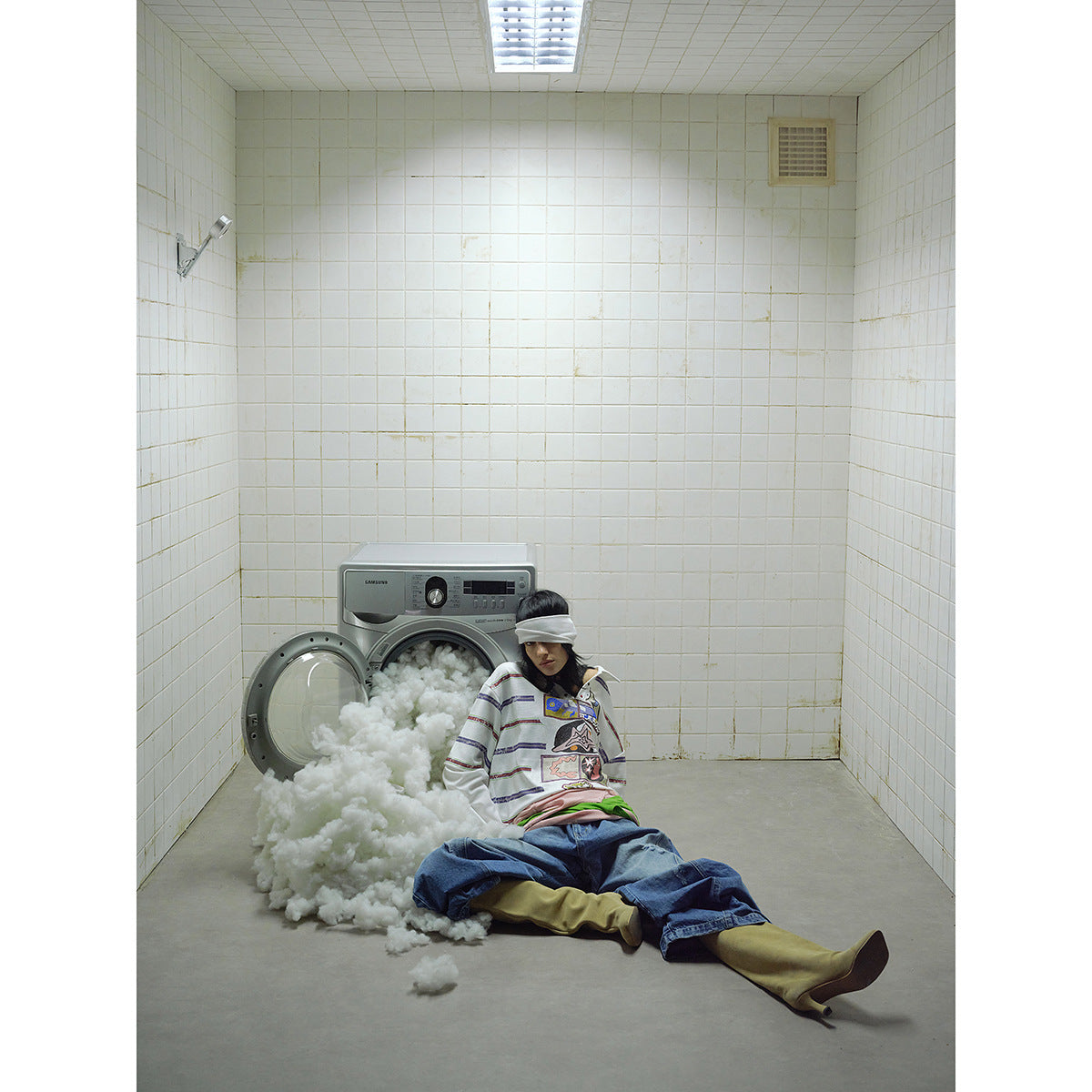 Wide scene of a model wearing a white striped graphic polo and baggy jeans beside an open washing machine.